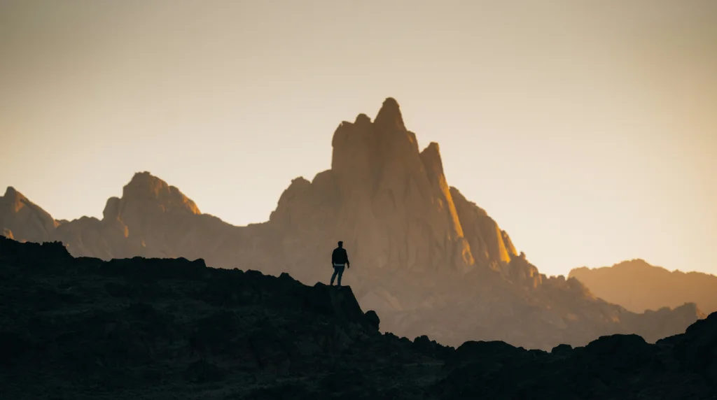 A man standing on a mountain from a distance, gazing at a range of mountains, symbolizing reflection and the journey of overcoming colon cancer